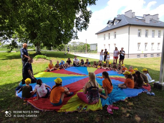 na zdjęciu policjantka wraz z dzielnicowym edukują dzieci które siedzą na kolorowej macie