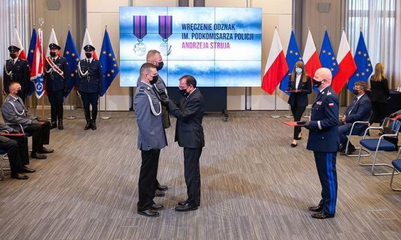 Fotografia przedstawiająca wręczenie odznaczeń. Minister Mariusz Kamiński przypina policjantowi odznaczenie. Z tyłu stoi Komendant Główny Policji.