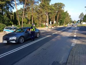 Na zdjęciu znajduje się policjant na jezdni stojący obok pojazdu osobowego podczas kontroli drogowej
