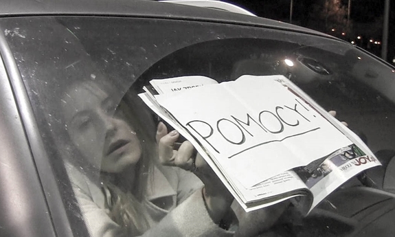 Biało czarna fotografia. Przednia szyba samochodu. W środku widać młodą kobietę, która trzyma biała kartkę z napisem w kolorze czarnym pomocy.