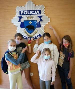 Zdjęcie kolorowe przedstawia grupę 5-ci dziewczynek które przebywaja w auli KMP-Przemyśl na spotkaniu profilaktycznym. Dziewczynki stoją pod ścianą na której umieszczona jest gwiazda policyjna.