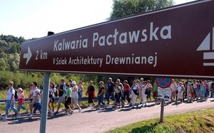 Zdjęcie kolorowe przedstawia znak drogowy informacyjny. Znak jest koloru niebieskiego (prostokątny) na znaku białmi literami wypisany jest napis:&quot;Kalwaria Pacławska&quot; pod znakiem widnieje drugi napis : &quot;2 km V szlak Architektury Drewnianej&quot;.
Za znakiem widoczna jest pielgrzymka która zmierza na odpust kalwaryjski