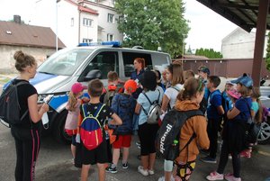 Zdjęcie kolorowe przedstawia parking KMP w Przemyślu. Na zdjęciu są widoczne dzieci które stoją przed policjantka umundurowaną. W tle widoczny oznakowany radiowóz.