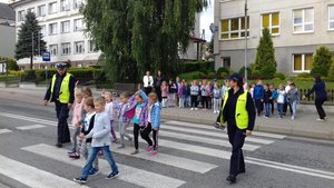Policjanci pokazują dzieciom, jak przechodzić przez przejście dla pieszych. Dwoje policjantów i grupka dzieci na przejściu
