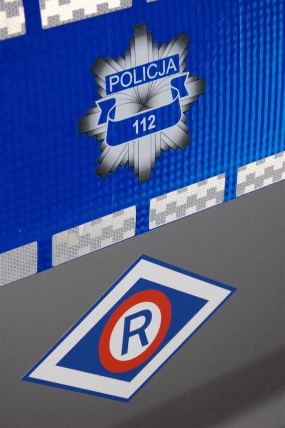 Na oznakowanym radiowozie Policji logo ruchu drogowego Duża litera &quot; R&quot; w kolorze granatowym. nad nią napis Policja i numer 112.