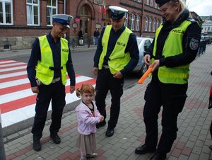 Zdjęcie kolorowe przedstawia trzech umundurowanych policjantów w kamizelkach odblaskowych którzy stoją obok małej dziewczynki i wręczają jej odblask