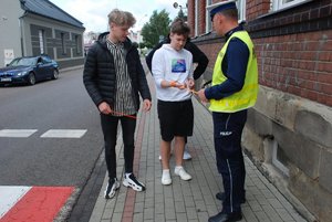 Zdjęcie kolorowe przedstawia ul. Kopernika w Przemyślu . Obok przejścia dla pieszych widoczny jest policjant umundurowany przy którym stoi trzech młodych chłopców. Policjant zakłada im opaski odblaskowe na ręce