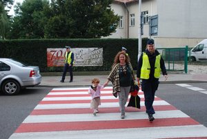 Zdjęcie kolorowe zrobione na ul. Kopernika w Przemyślu przy SOSW .Na zdjęciu widoczni są policjanci oraz policjantka która przeprowadza dziecko na pasach . Zdjęcie z akcji Bezpieczna droga do szkoły