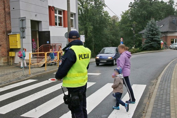 policjant w kamizelce odblaskowej stojący przy przejściu dla pieszych, przez które przeprowadzane jest dziecko