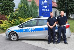 Policjanci w umundurowaniu służbowym w tle radiowóz za nimi pylon Komendy Powiatowej Policji w Lubaczowie dalej krzewy i drzewa.