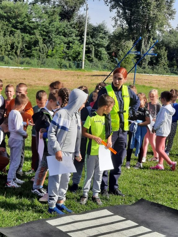 Zdjęcie kolorowe wykonane w porze dziennej przedstawia boisko szkolne SP w Nehrybce. Na boisku widoczne są dzieci oraz policjantka umundurowana, która w ramach akcji &quot;Bezpieczna droga do szkoły&quot;, wygłasza prelekcje oraz uczy dzieci przechodzenia przez nieoznakowane przejście dla pieszych.