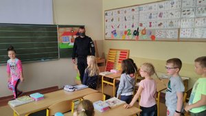 Zdjęcie kolorowe wykonane w porze dziennej , na zdjęciu widoczna jest klasa szkolna w której przebywają dzieci oraz policjant umundurowany, który w ramach akcji &quot;Bezpieczna droga do szkoły &quot; przeprowadza zajęcia.