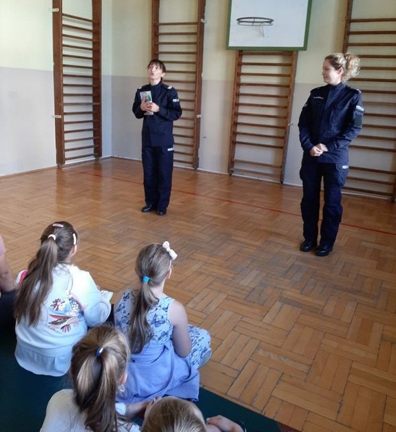 Umundurowane policjantki prowadza zajęcia dla dzieci. Uczniowie siedzą w sali gimnastycznej przodem do funkcjonariuszek.
