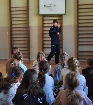 Umundurowane policjantki prowadza zajęcia dla dzieci. Uczniowie siedzą w sali gimnastycznej przodem do funkcjonariuszek.
