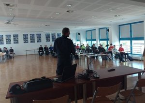 widok na salę, przy ławkach siedzą uczniowie, przed nimi stoi policjant