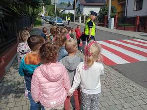 Zdjęcie kolorowe wykonane w porze dziennej. Na zdjęciu widoczne są dzieci oraz policjanci, którzy udają sie z dziećmi na przejście dla pieszych aby uczyć ich jak w sposób bezpieczny przechodzić przez jezdnię