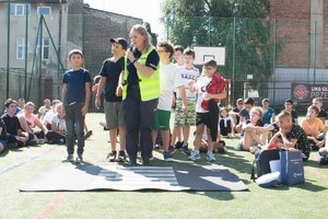 Zdjęcie kolorowe wykonane w porze dziennej. Przedstawia boisko szkolne na którym przebywają dzieci i znajdują się obok policjantki umundurowanej , która przeprowadza prelekcję na temat bezpieczeństwa