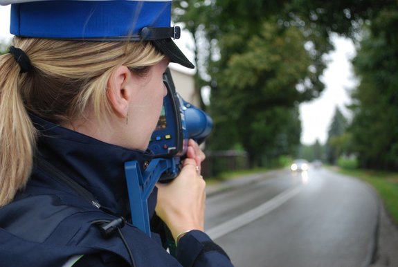 Policjantka ruchu drogowego stojąc przy drodze mierzy prędkość laserowym miernikiem prędkości.