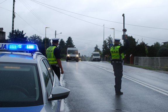 Policjanci drogówki zatrzymujący na drodze kierowce busa do kontroli. Na pierwszym planie radiowóz z włączonymi sygnałami świetlnymi.
