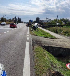 Zdjęcie kolorowe, wykonane w porze dziennej w miejscowości Orły . Zdjęcie przedstawia samochód m – ki volkswagen transit koloru białego, który jest zaparkowany w rowie.  Po lewej stronie samochodu widoczna jest koparka o małych gabarytach w kolorze żółtym.