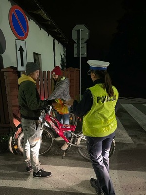 Na zdjęciu znajduje się policjantka wręczająca elementy odblaskowe rowerzystom w porze wieczorowo-nocnej