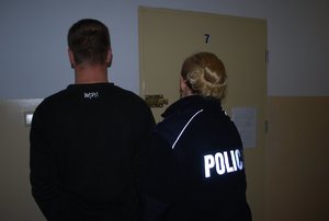 na zdjęciu umundurowana policjantka wprowadza do celi w policyjnym areszcie zatrzymanego w związku z rozbojem mężczyznę. Obydwoje stoją tyłem przed drzwiami celi.