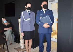 Policjantka i policjant stoją twarzą w stronę obiektywu. W tle kobieta siedząca na krześle.