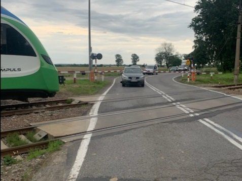 Zdjęcie poglądowe - kierujący samochodem osobowym stoi pomiędzy opuszczonym szlabanem na przejeździe kolejowym a jadącym torami pociągiem