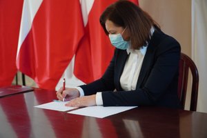 Wojewoda Ewa Leniart podpisuje dokument porozumienia. W tle biało-czerwone flagi