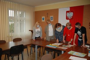 . Zdjęcie kolorowe wykonane w porze dziennej przedstawia aulę Komendy miejskiej Policji w Przemyślu.
Na fotografii widoczne sa stoły na których leżą prace rysunkowe dzieci i młodzieży które biorą udział w konkursie plastycznym „Bezpieczne wakacje 2021”. Nad pracami stoi 5 osób -komisja konkursu .