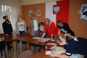 Zdjęcie kolorowe wykonane w porze dziennej przedstawia aulę Komendy miejskiej Policji w Przemyślu.
Na fotografii widoczne sa stoły na których leżą prace rysunkowe dzieci i młodzieży które biorą udział w konkursie plastycznym „Bezpieczne wakacje 2021”. Nad pracami stoi 7 osób -komisja konkursu .