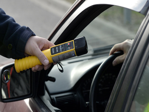 Zdjęcie kolorowe przedstawia urządzenie do pomiaru alkoholu w wydychanym powietrza „alcosensor”, który jest koloru żółtego z czarnym ustnikiem. Urządzenie jest podawane  przez okno pojazdu osobowego.