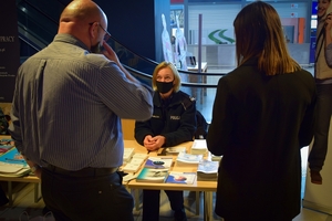 Policyjne stoisku w Porcie Lotniczym Jasionka - Rzeszów. Funkcjonariusze siedzący przy stole z ulotkami rozmawiają z zainteresowanymi osobami.