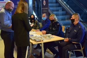 Policyjne stoisku w Porcie Lotniczym Jasionka - Rzeszów. Funkcjonariusze siedzący przy stole z ulotkami rozmawiają z zainteresowanymi osobami.