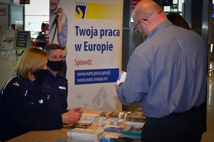 Policyjne stoisku w Porcie Lotniczym Jasionka - Rzeszów. Funkcjonariusze siedzący przy stole z ulotkami rozmawiają z zainteresowanymi osobami.