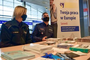 Policyjne stoisku w Porcie Lotniczym Jasionka - Rzeszów. Funkcjonariusze siedzący przy stole z ulotkami rozmawiają z zainteresowanymi osobami.