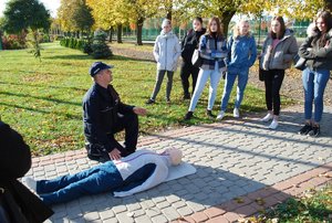 Na zdjęciu, na placu przed szkoła podkom Jacek Siek podczas prelekcji o zasadach udzielania pierwszej pomocy przedmedycznej. Policjant klęczy nad fantomem, wokół niego stoją słuchające prelekcji uczennice.