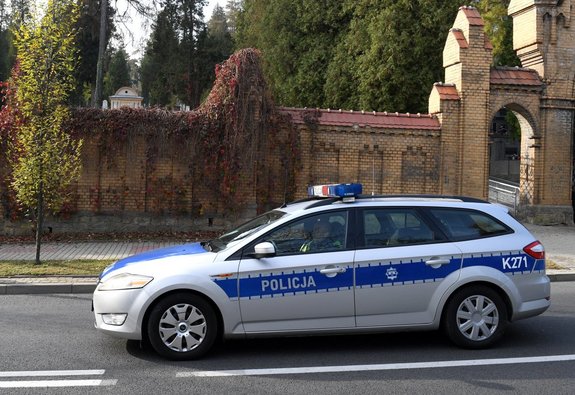 Zdjęcie wykonane w porze dziennej przedstawia cmentarz główny w Przemyślu przy ul. Słowackiego. Przed cmentarzem zaparkowany jest policyjny pojazd uprzywilejowany marki Kia. Radiowóz jest w kolorze srebrnym z niebieskimi pasami na środku pojazdu z napisem POLICJA w białym kolorze.
