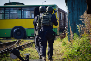Uzbrojeni policyjni kontrterroryści schowani za tarczą idą w kierunku autobusu. Policjanci widoczni od tyłu