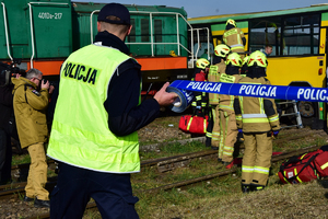 Na środku w tle lokomotywa i autobus - strażacy udzielają pomocy poszkodowanym. Na pierwszym planie policjant rozwija niebieską taśmę z napisem POLICJA