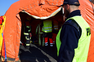 Służby medyczne w pomarańczowym namiocie udzielają pomocy poszkodowanym. Na pierwszym planie po prawej policjant w kamizelce odblaskowej