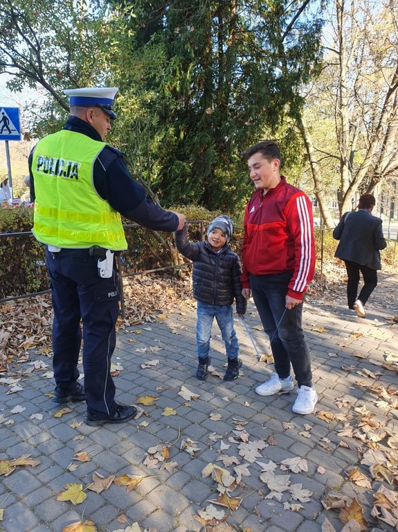 Zdjęcie kolorowe wykonane w porze dziennej przedstawia cmentarz w Przemyślu przy ul. Słowackiego. Przed cmentarzem widoczny policjant z kobieta i małym chłopcem. Policjant wręcza chłopcu odblaski