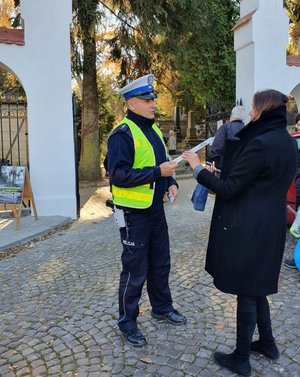 Zdjęcie kolorowe wykonane w porze dziennej przedstawia cmentarz w Przemyślu przy ul. Słowackiego. Przed cmentarzem widoczny policjant z kobieta . Policjant wręcza kobiecie odblaski