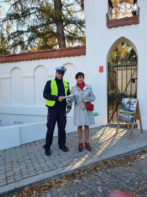 Zdjęcie kolorowe wykonane w porze dziennej przedstawia cmentarz w Przemyślu przy ul. Słowackiego. Przed cmentarzem widoczny policjant z kobieta i małym chłopcem. Policjant wręcza chłopcu odblaski