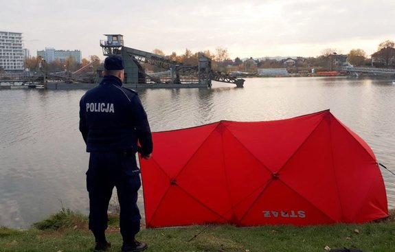 Umundurowany policjant stojący na brzegu zalewu nad Wisłokiem. Przed nim stoi czerwony parawan, w tle rzeka