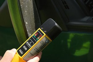 Zdjęcie kolorowe przedstawia urządzenie do badania zawartości alkoholu w  wydychanym powietrzu. Urządzenie „Alcoblov” jest w kolorze żółtym a ustnik jest koloru czarnego.