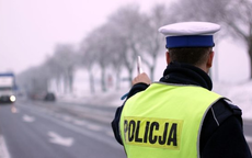 Zdjęcie kolorowe wykonane w porze dziennej przedstawia policjanta ruchu drogowego stojącego tyłem . Policjant stoi na drodze w ręku trzyma tarczę do zatrzymywania pojazdów z przeciwka widoczny samochód, który porusza się w stronę policjanta.