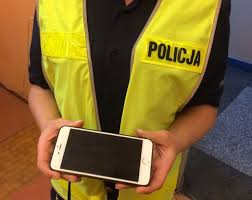 Zdjęcie kolorowe przedstawia policjantkę w ubraniu cywilnym z założoną kamizelka odblaskowa koloru żółtego z czarnym napisem POLICJA po prawej stronie, która w reku trzyma telefon komórkowy.