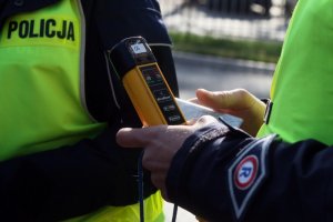 Na zdjęciu widoczni są częściowo dwaj policjanci ruchu drogowego umundurowani i posiadający na sobie kamizelki odblaskowe. Jeden z policjantów prawej strony trzyma w reku urządzenie do pomiaru alkoholu w wydychanym powietrzu „Alboblov” koloru żółtego z czarna końcówką.