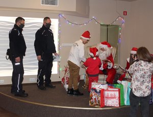 Na zdjęciu widoczny Mikołaj, pomocnica Mikołaja, dwaj umundurowani policjanci stojący na scenie wśród prezentów i dzieci odbierające prezent.